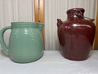 Green glazed pottery pitcher with ridged texture and chip on rim next to red pottery jug with metal handle, shown together.