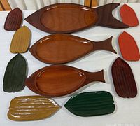 Top down view showing three polished mahogany fish-shaped plates of different sizes, and six leaf-shaped wooden dishes in various colors arranged around them.
