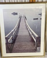 Framed black and white photographic print depicting a wooden pier extending into a lake with small boats floating in the distance. The image is crisp, matte with a white border and framed in a thin gold frame.