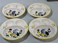 Four ceramic egg plates with three egg holders each and central rooster and chick decoration, showing overall condition and paint wear.