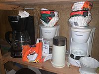 Three Black & Decker coffee makers lined up on a wooden shelf with paper coffee filters stacked on them and a white Braun coffee grinder with clear plastic top container on shelf.