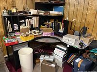 Overview of basement office corner showing black shelving unit, desk with curved glass top, bags, boxes, binders, and stationery supplies scattered and stacked.