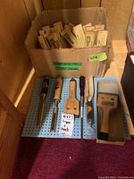 Lot of chisels and scraper displayed on blue pegboard with cardboard box of replacement blades behind