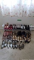 Wide view showing 17 pairs of women's high heel shoes arranged around a white metal shoe rack with two levels, placed on carpet in front of window with white floral curtains.