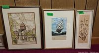 Three framed items in a row: the 16x13 riverside town painting on the left, the 15x12 tall ship painting in the middle, and the 15x6 dried flower art on the right.
