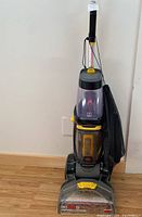 Front view of the Bissell Revolution upright carpet cleaner showing gray, black, and yellow plastic body, clear dirt tank, handle, and yellow controls.