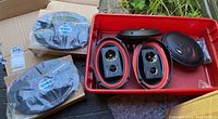 View of the lot with multiple speaker units arranged in a red plastic bin and adjacent to a cardboard box outdoors. Shows two pairs of Scosche 6x9 speakers in packaging and other speakers unpackaged.