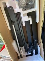 Photo of disassembled black metal desk frame parts laid in foam packaging inside a cardboard box with included power adapter and cables.