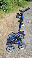 Blue metal folding golf push cart standing upright in partially folded position on gravel with grass in background.