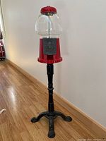 Full view of red gumball machine on tall black metal stand against wall corner on wooden floor.