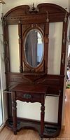 Full view of dark-stained wooden hall tree with oval mirror, drawer, brass hooks and umbrella stands