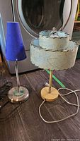 Two shaded desk lamps side by side on floor, one with blue conical shade and silver base, other with light blue double-tiered shade and wooden base