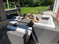 Overall assembly of five wicker seating modules arranged around a central glass-top wicker table on an exterior deck adjacent to a wood pile and grass lawn.