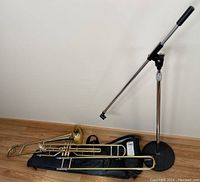 Two trombones with valves lying on a damaged soft carrying case beside an adjustable microphone stand with round black base.