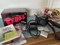 Canon EOS Rebel T2i camera body with original box, charger, 2 batteries, AV cable, manuals, and camera strap on a table.