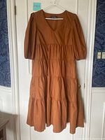Full front view of a light brown dress on hanger against closed door showing the tiered design, V-neck, and puff sleeves
