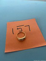 Gold ring placed on orange paper with the number 157 written on it, showing front view of ring.