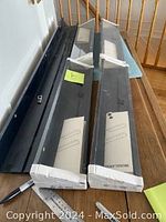 Four IKEA Mosslanda picture ledges laid on the floor next to a staircase, all black with packaging showing the product label. One long ledge marked 115, three shorter ones marked 55.