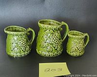 Front view of three green Japanese ceramic measuring cups in 1 cup, 1/2 cup, 1/4 cup sizes with embossed grape and leaf designs.