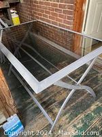 Full angled view of rectangular patio table with black metal frame and transparent tempered glass top on a wooden floor next to brick wall.
