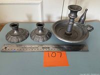 Two pewter candlesticks with flared stepped bases and one pewter chamberstick with snuffer on a surface with ruler and orange lot number card