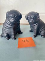 Two matte black weighted pug dog figurines shown side by side on a surface with a note labeled 93.