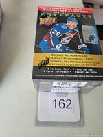 Photo of sealed 2021-2022 Upper Deck Artifacts Hockey blaster box with box artwork featuring a player in blue helmet and jersey, unopened packaging with promotional text about autograph and game-worn memorabilia cards.