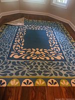 Wide view of the rug showing the full laurel border design and central blue area laid out on wood floor in a room with windows.