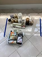 Lot of assorted candles, glass holders, soaps, lotion tubes, chrome hook and scale arranged on tiled floor.