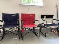 Two Freestyle Rocker brand folding rocking chairs, navy blue and red, alongside a black director's chair with a side table attachment on the armrest. Shown on a garage floor under a window.