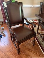 Close-up of a captain chair showing dark brown leather upholstery, nailhead trim, carved wooden arms, and turned legs on hardwood floor
