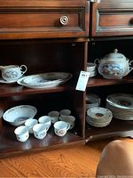 Wide view showing multiple porcelain items including cups, saucers, serving platters, bowls, gravy boat with cover, and plates arranged on wooden shelving