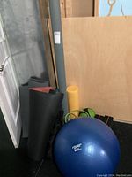 Photo showing a blue 65 cm Fitness Gear exercise ball, black rolled mats, yellow foam mat, and various workout accessories stored near a door and a wooden board.