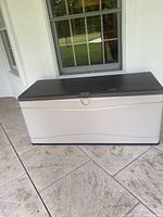 Front view of the rectangular vinyl outdoor deck box with beige body and dark gray lid showing the handle and some dirt marks.