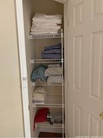 Shelf with folded white sheets on top, blue and green sheets or pillowcases, beige/light towels or pillow shams lower down, and two white pillows. Bottom shelf contains red and gray washcloths or small towels.