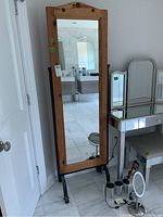 Full-length wood framed cheval mirror with metal base stand, capturing front view of the mirror reflecting bathroom and part of vanity desk with vanity mirror in the background.