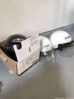 Three HJC motorbike helmets: black in box, two white with visors.