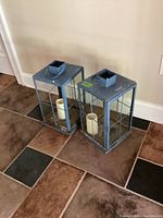 Two metal and glass lanterns on tiled floor showing front and side views with flameless candles inside.