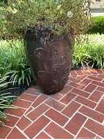 Full view of the large dark brown ceramic planter placed outdoors on a brick patio, showing embossed floral design and glossy finish with some weather exposure.