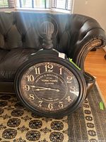 Full view of the large round wall clock on a patterned rug in front of a leather armchair. The clock is black with cream colored numbers and an attached handle on top mimicking a pocket watch.