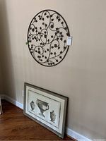 Round metal decal with vine and leaf details hanging on wall above framed classical urn print leaning against wall
