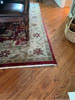 Close-up image capturing the border design and texture of the wool area rug showing red and beige floral motifs along the edges on the hardwood floor.
