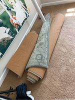 Two rolled rugs on carpet floor near wall; one is 3x5 size with a visible floral border pattern, the other larger and rolled with beige non-slip mats visible.