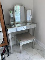 Front view of mirrored vanity with tri-fold mirror and upholstered bench set against white wall on marble flooring