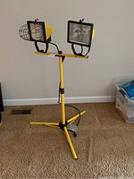 Standing dual head work light with yellow metal frame, two rectangular heads with one missing bulb in each, tripod base on carpet.