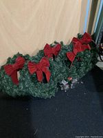 Five green artificial Christmas wreaths lined up, each decorated with large red velvet bows, on a dark floor against a beige wall.