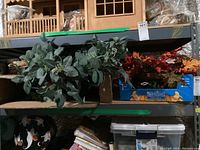 Photo of two main wreaths on shelving, a green eucalyptus wreath and a fall wreath with red and orange leaves, along with boxes containing wreath components and hangers.