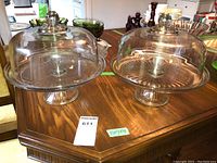 Two identical clear glass domed pedestal cake stands on a wooden table, showcasing the overall size and design.