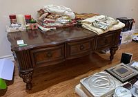 Wood coffee table with three drawers, metal ring pull hardware, decorative carved front and legs. Various textile items and small objects on top are not included.