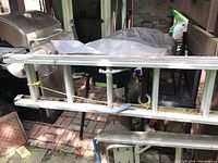 Side view of the aluminum extension ladder showing rungs and side rails secured with a yellow cord, placed on a black metal surface outdoors with a grill in the background.